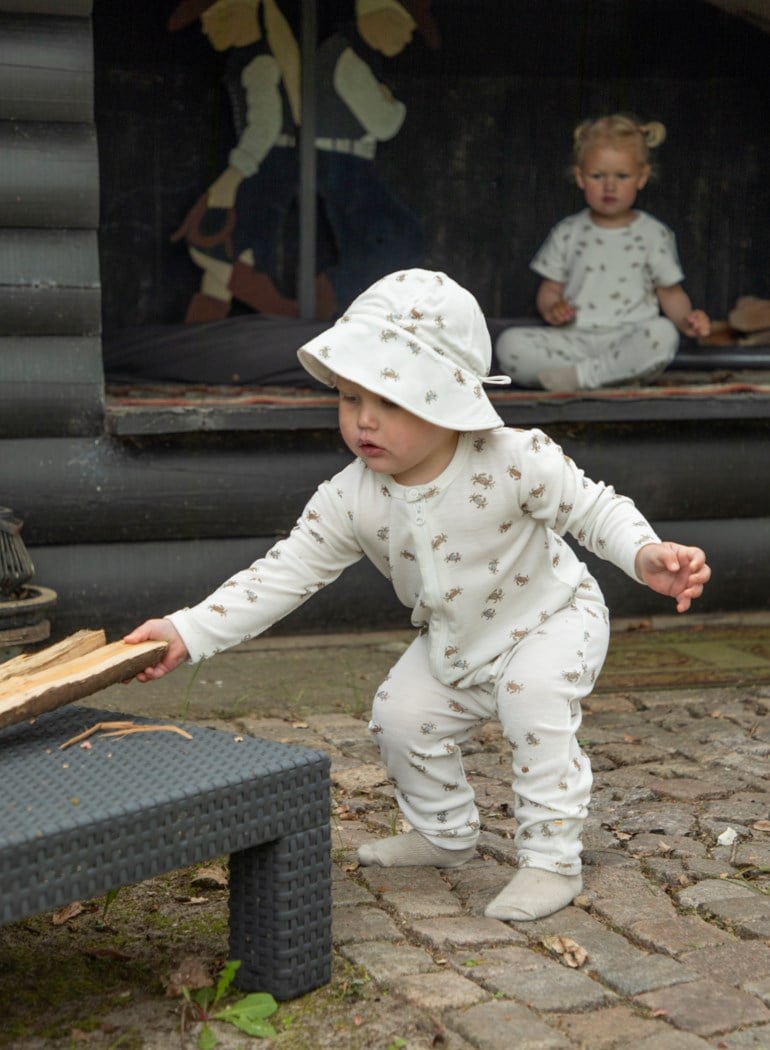 Baby i beige krabbemønstret glidelåsdrakt og solhatt, holder ved; annen baby har matchende todelt sett.