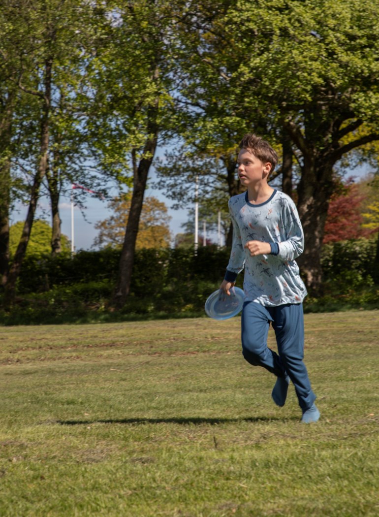 Barn i lyseblå mønstret langermet topp og matchende bukser, holder en klarblå frisbee.