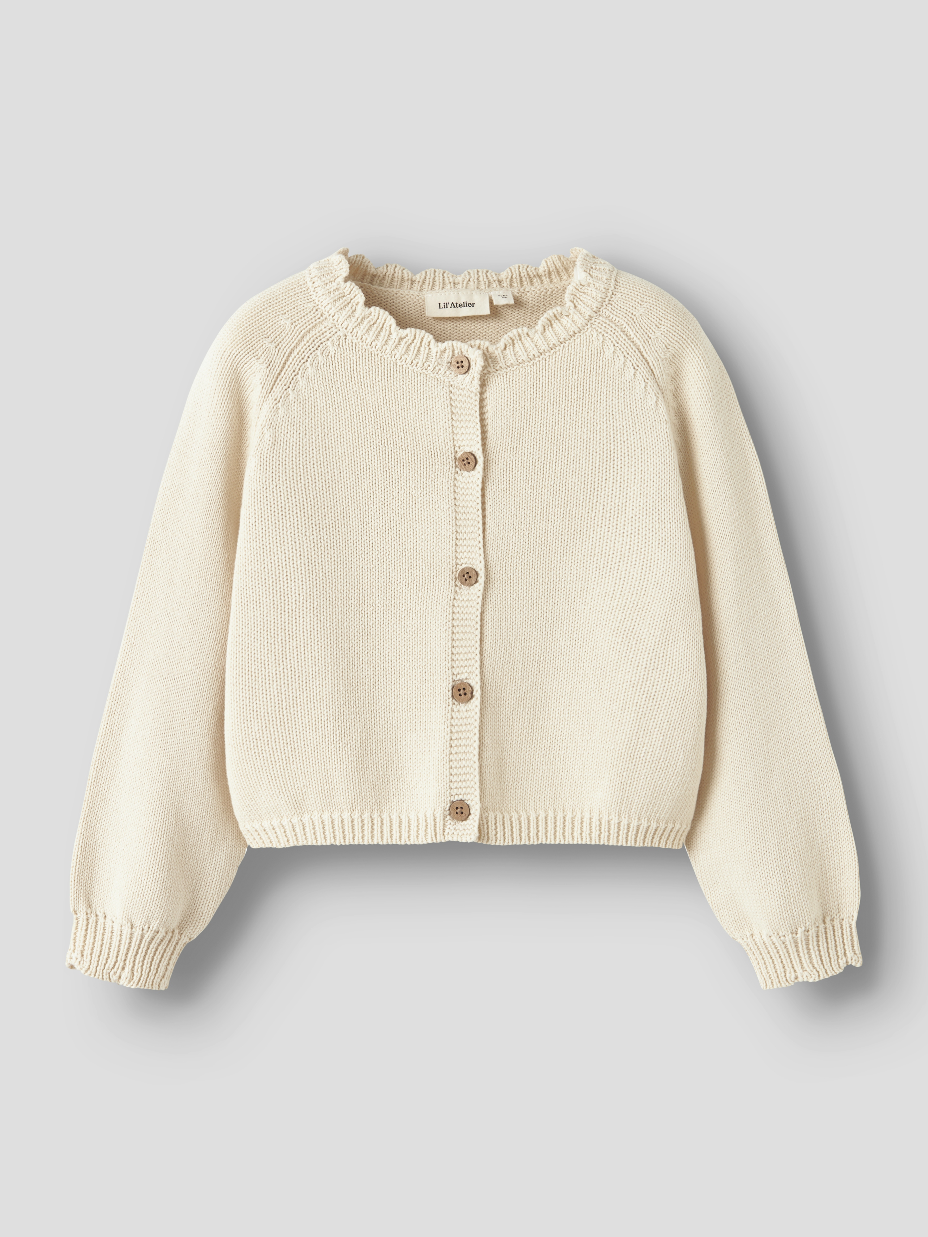 Kremfarget strikket barnegenser med volangkrage, raglanermer, ribbestrikkede mansjetter og knappelukking.