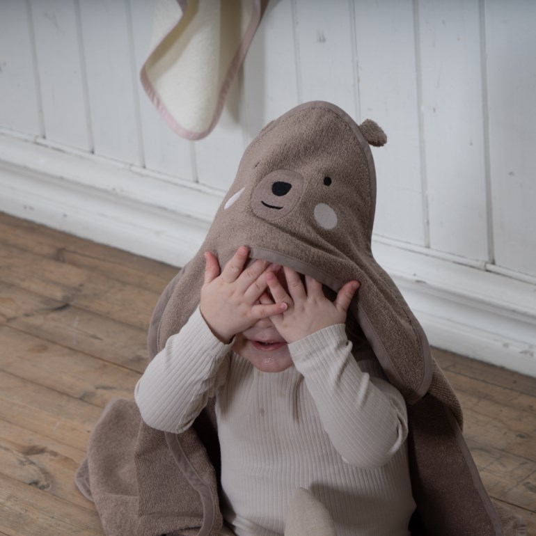 Baby iført brunt bjørnehettehåndkle over lys beige ribbet langermet topp, dekker ansiktet.