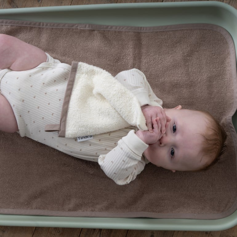 Baby holder kremfarget frottévaskeklut, har på kremfarget ribbet body med hjerter på brunt stelleunderlag.