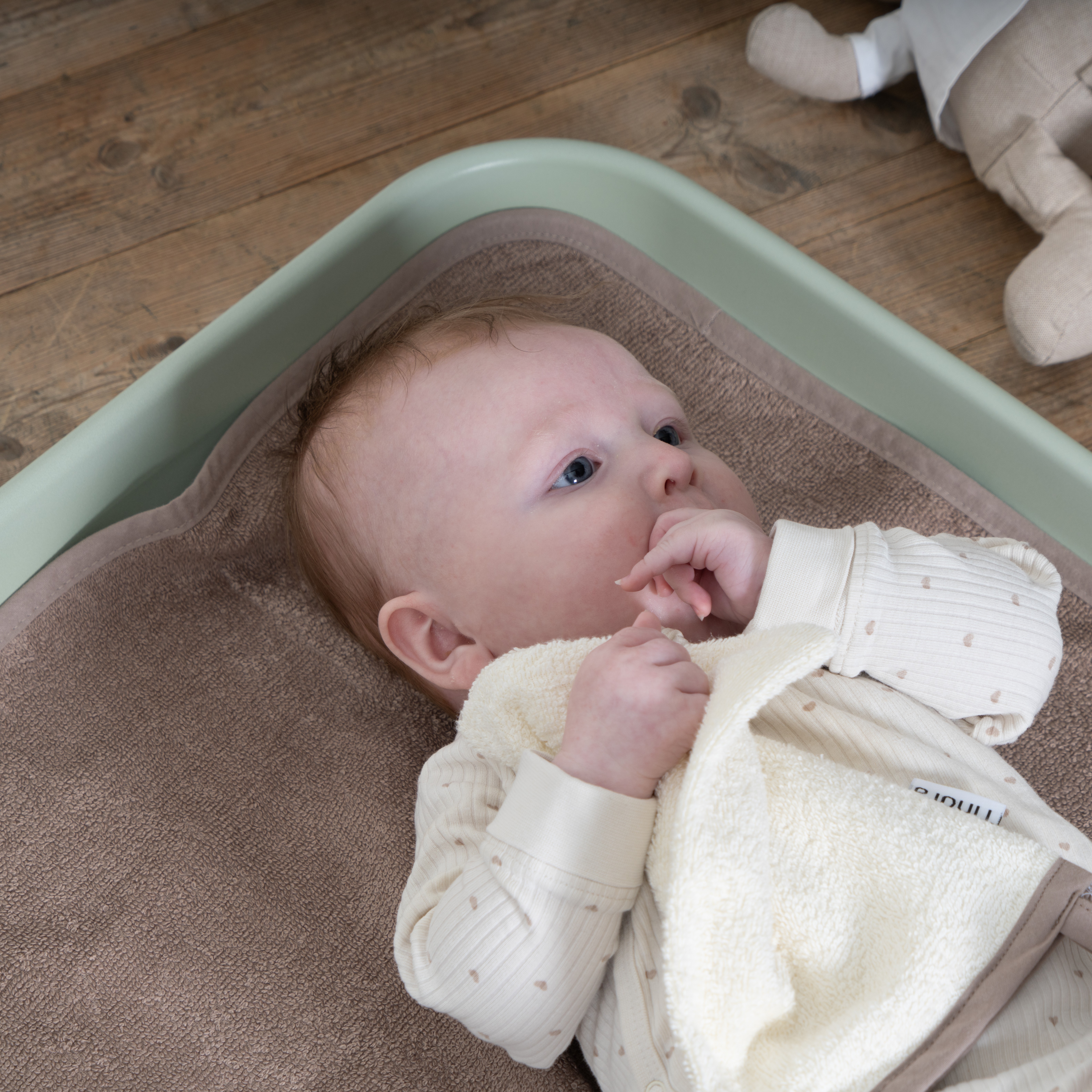 Baby i kremhvit ribbet body med hjertemønster, holder frottehåndkle, på brun stellematte i grønn balje.