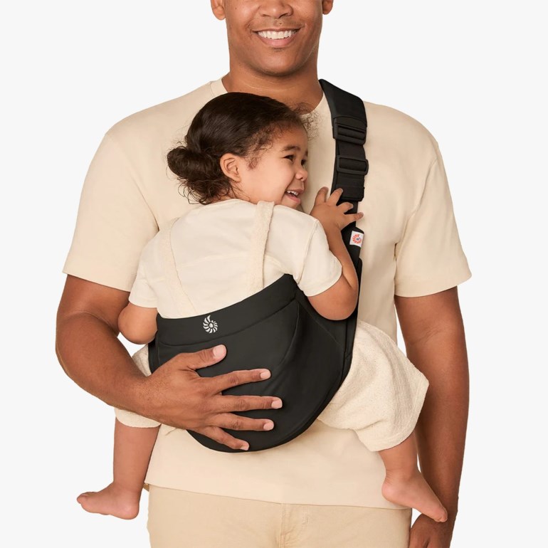 Svart bæresele med hvit logo, båret av en mann som bærer et smilende barn i kremfarget antrekk.