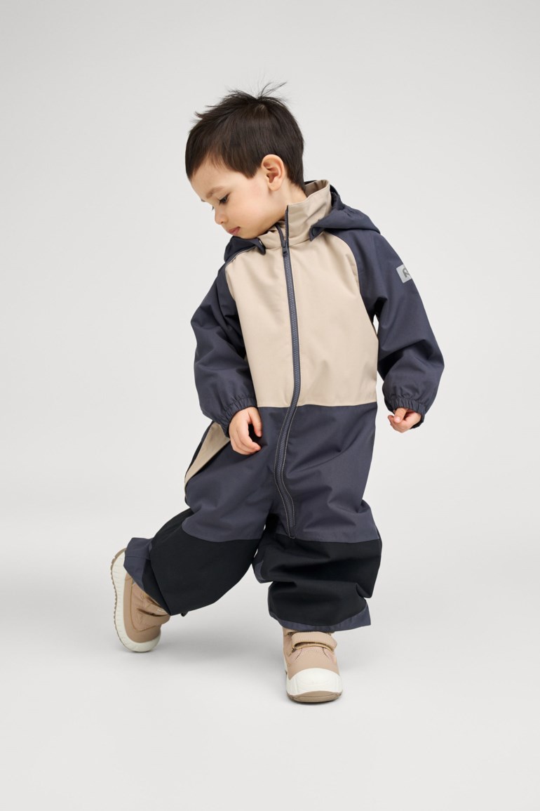 Barn iført en beige og mørkeblå hette vanntett heldress med svarte forsterkede knær og lysebrune sko.
