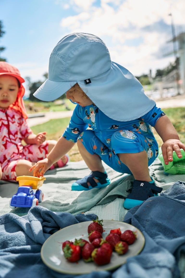Lys blå barnesolhatt med nakkeklaff, blåmønstret baderdrakt og mørkeblå sandaler. Annet barn i rød hatt.