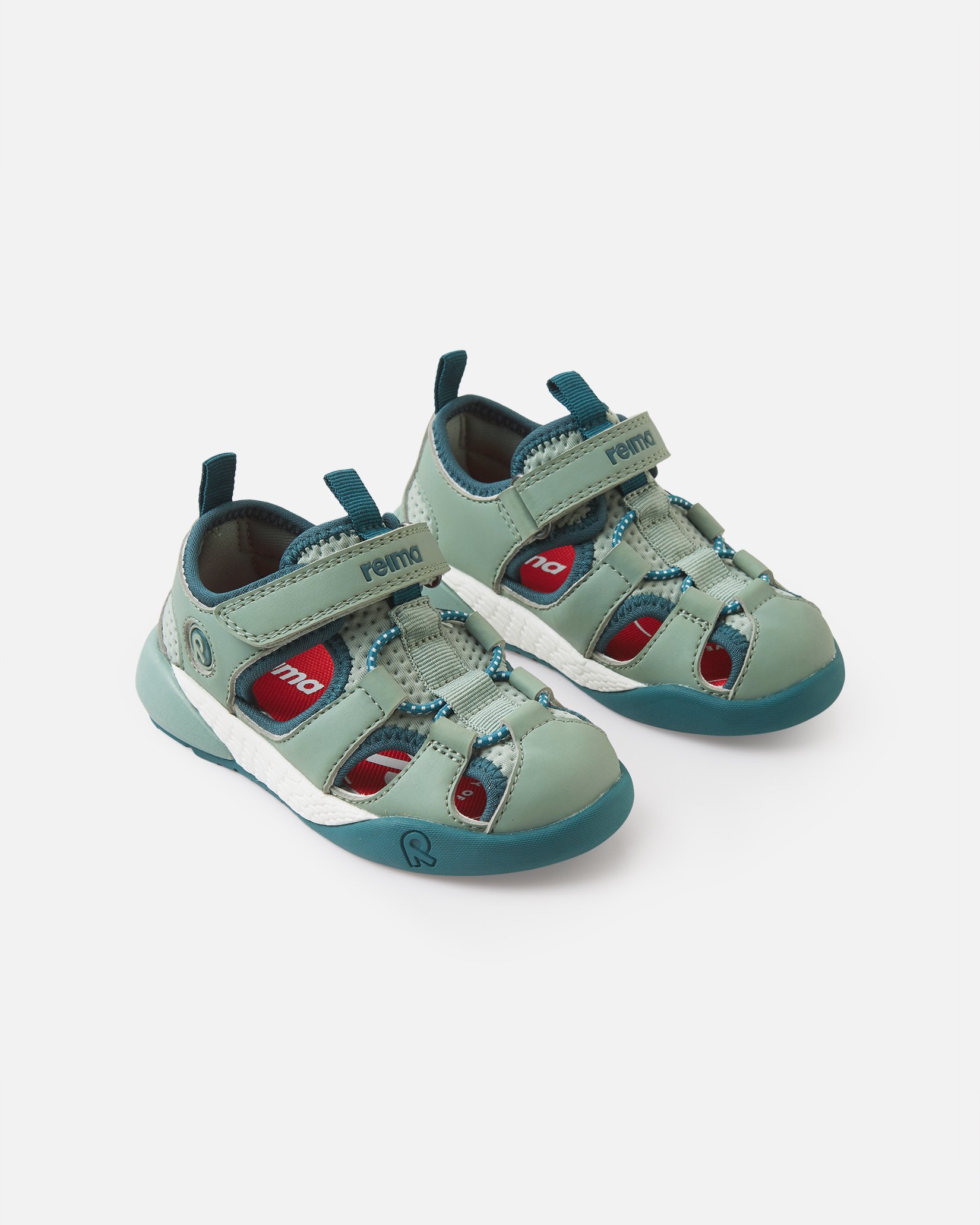 Et par grågrønne og mørkeblå barnesandaler med lukket tå, røde detaljer og hvite såler, forfra.