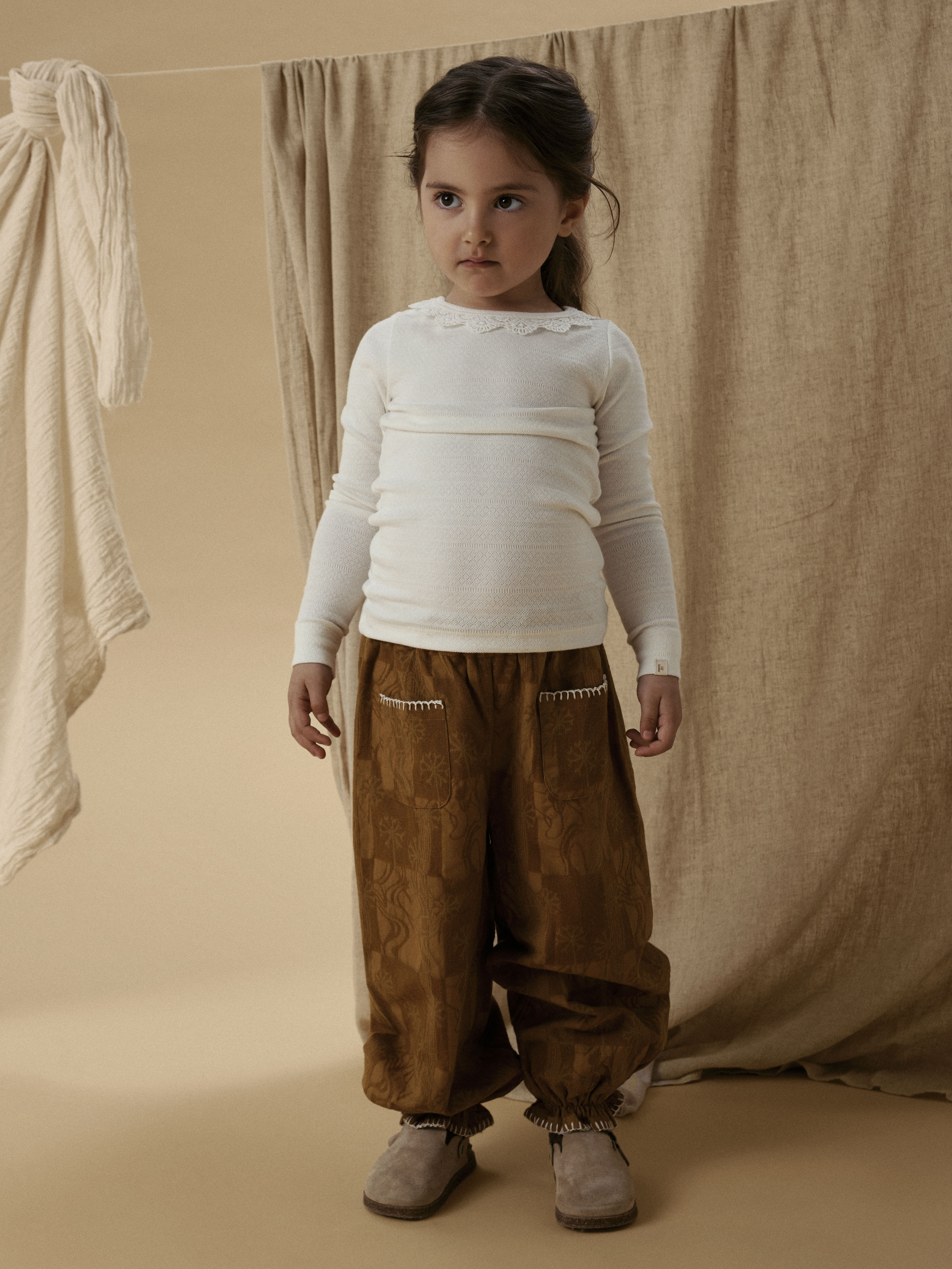 Barn i brune mønstrede vide bukser med hvit kant, hvit ribbet langermet topp og beige sko.