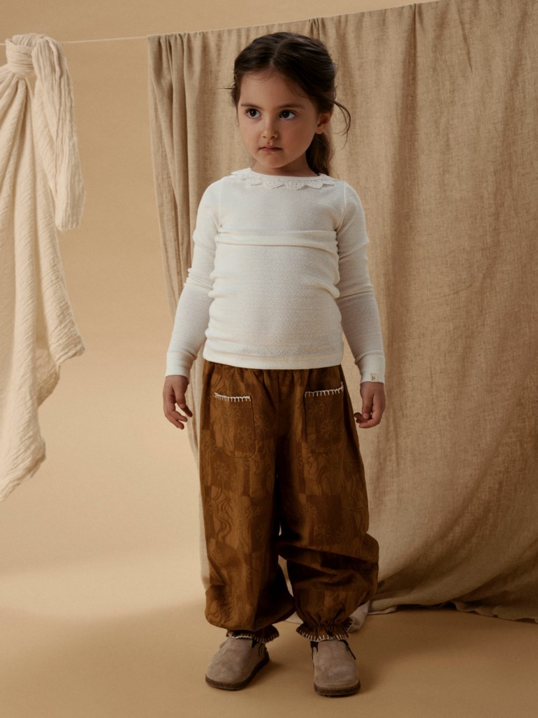 Barn i brune mønstrede vide bukser med hvit kant, hvit ribbet langermet topp og beige sko.