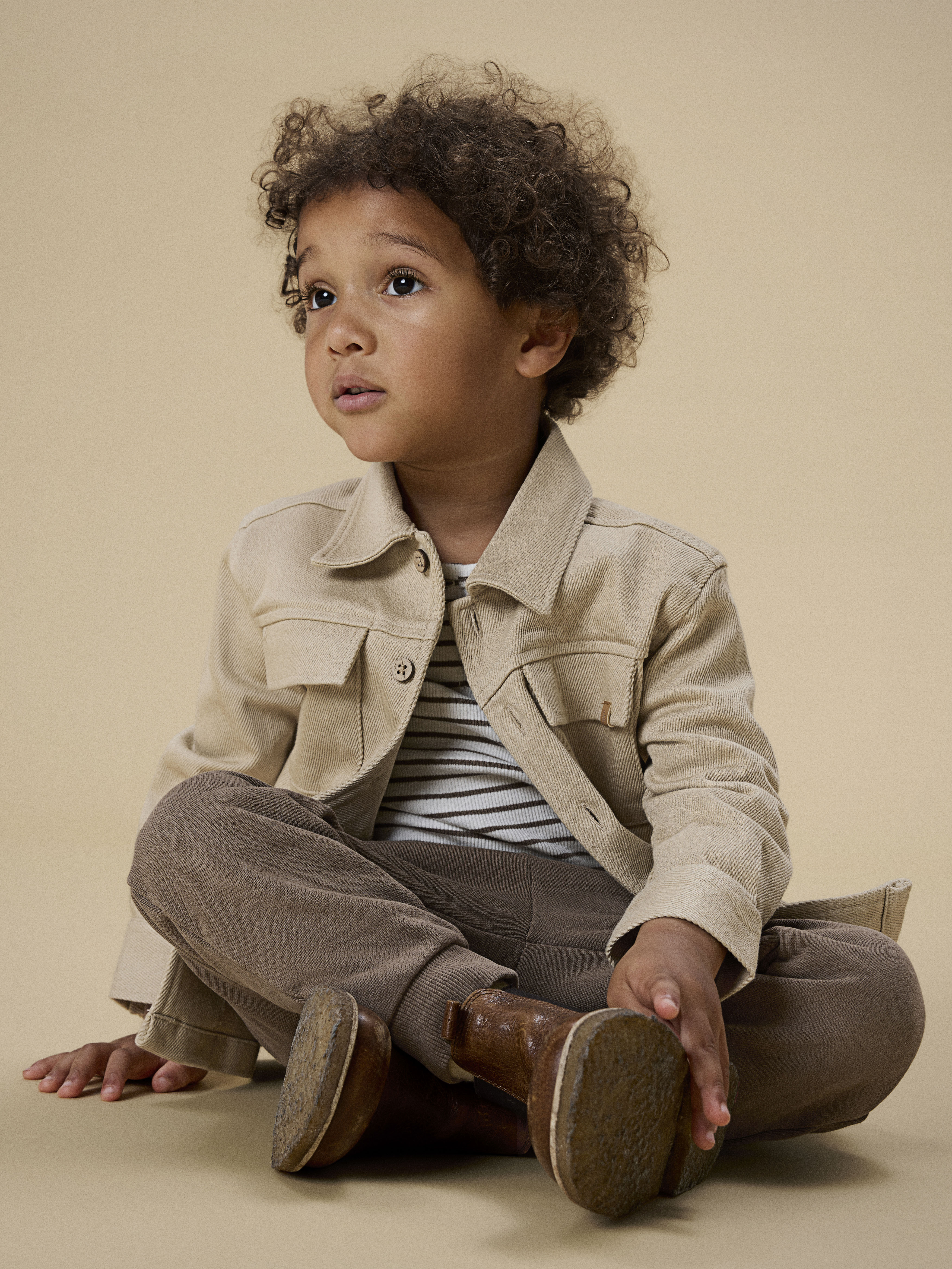 Barnets beige kordfløyelsjakke med brystlommer over en stripete t-skjorte, brune joggebukser og brune sko.