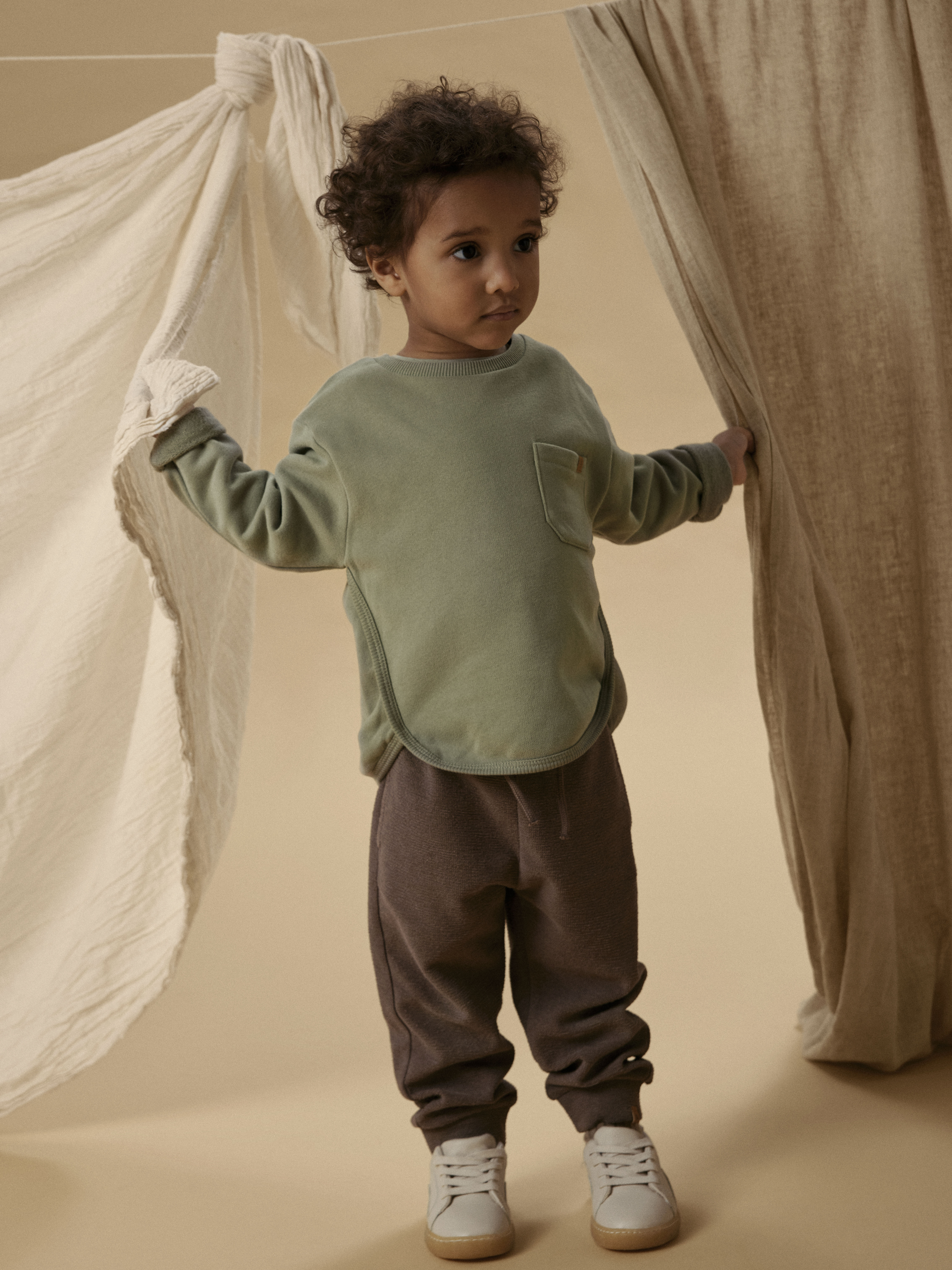 Kids grønn langermet genser med brystlomme, mørkebrune joggebukser og hvite joggesko.