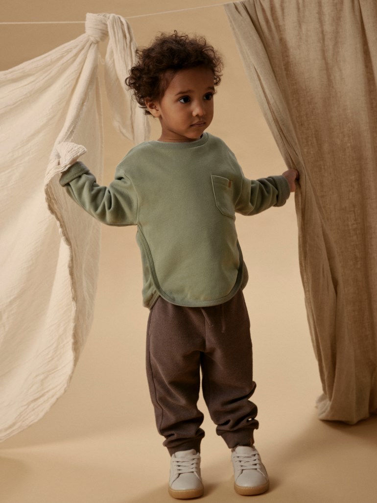 Kids grønn langermet genser med brystlomme, mørkebrune joggebukser og hvite joggesko.