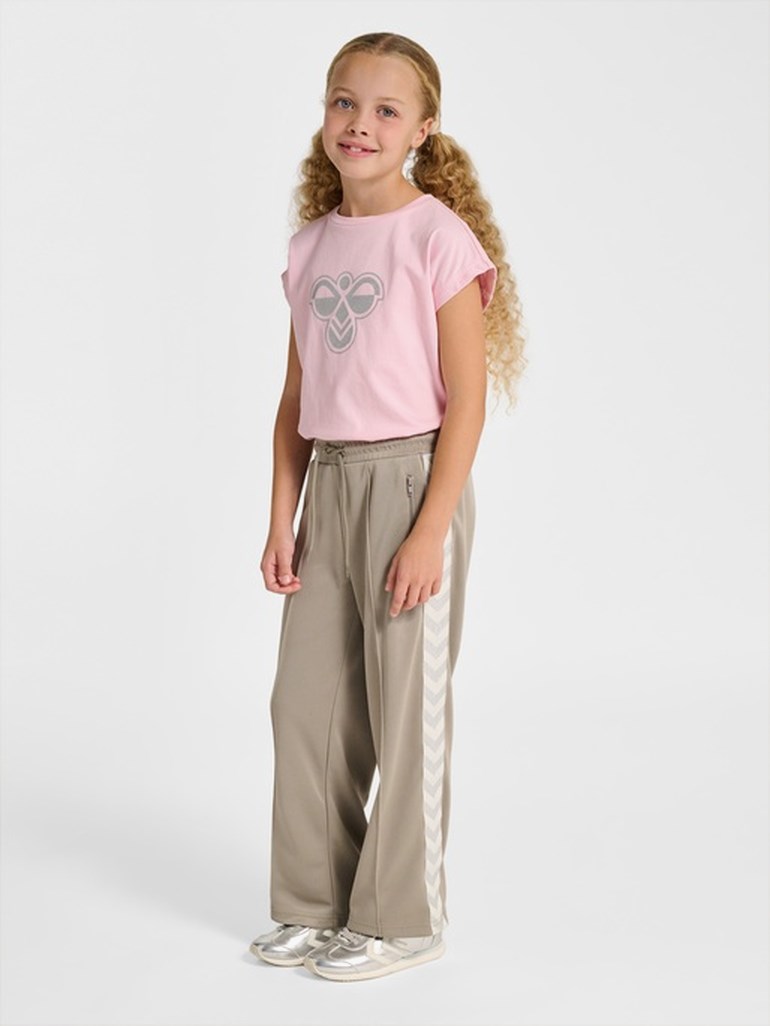 Jente iført en lys rosa kortermet t-skjorte med grå logo, lysebrune vide treningsbukser og sølvsneakers.