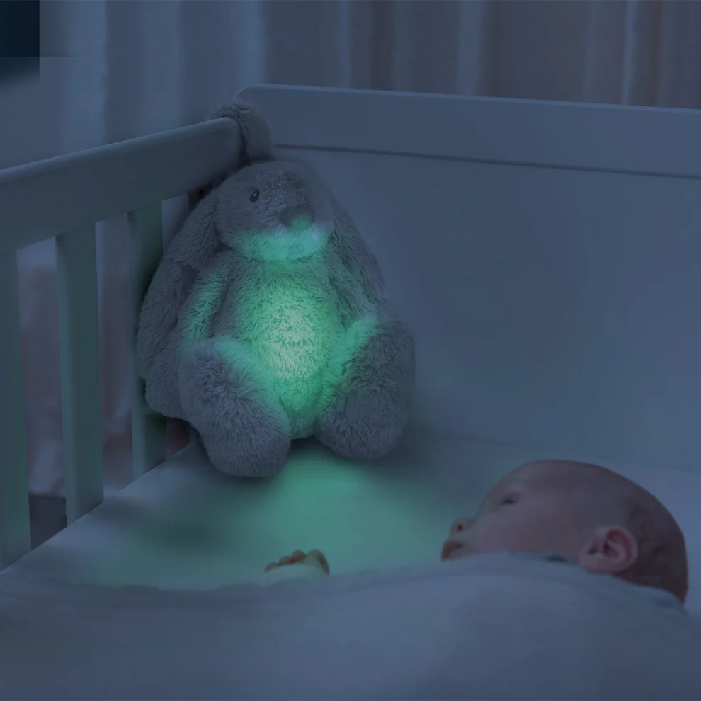 En grå lysende kaninleke sitter i en hvit babyseng med en sovende baby.