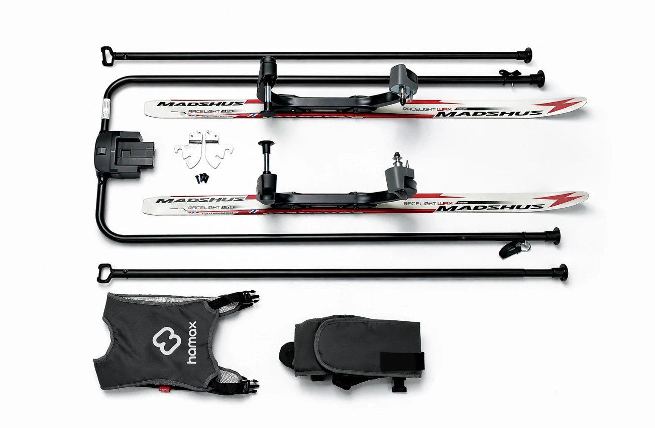 Demontert skipulksystem for barn med to hvite ski, svart metallramme, svart sele og oppbevaringsveske.