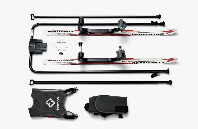 Demontert skipulksystem for barn med to hvite ski, svart metallramme, svart sele og oppbevaringsveske.