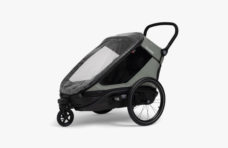 En grå og svart Hamax sykkelvogn for barn med klart regntrekk og hjul, sett fra siden.