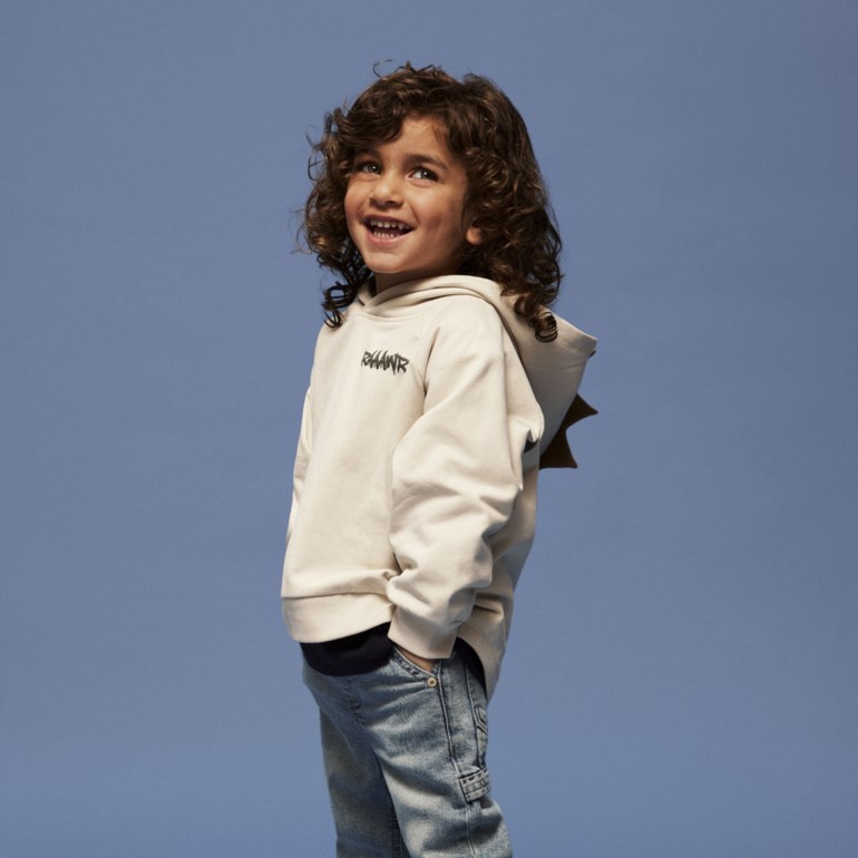 Barn bruker en lys beige hettegenser med dinosaurpigger og 'RRAWR' trykk, lyseblå jeans og beige sneakers.