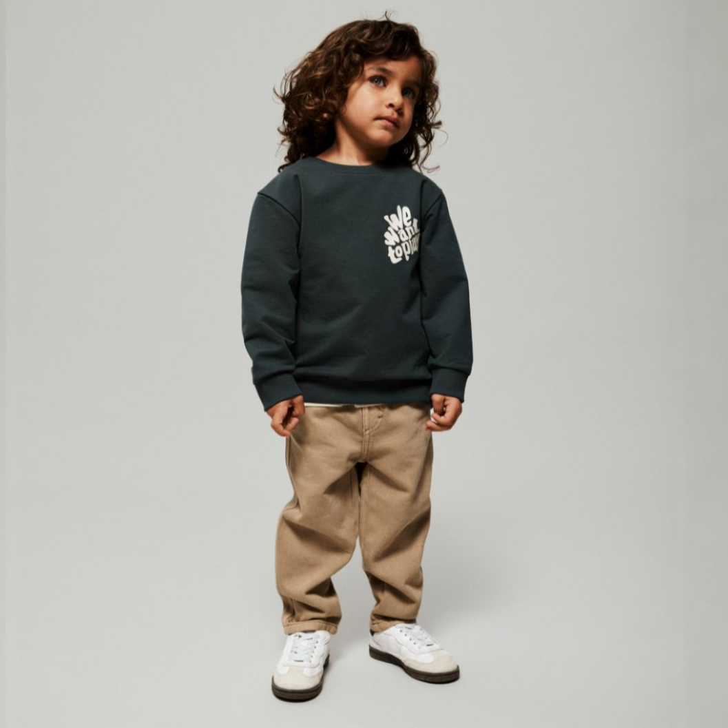 Et barn i en mørkegrønn sweatshirt med trykk, lysebrune bukser og hvite og brune joggesko, forfra.
