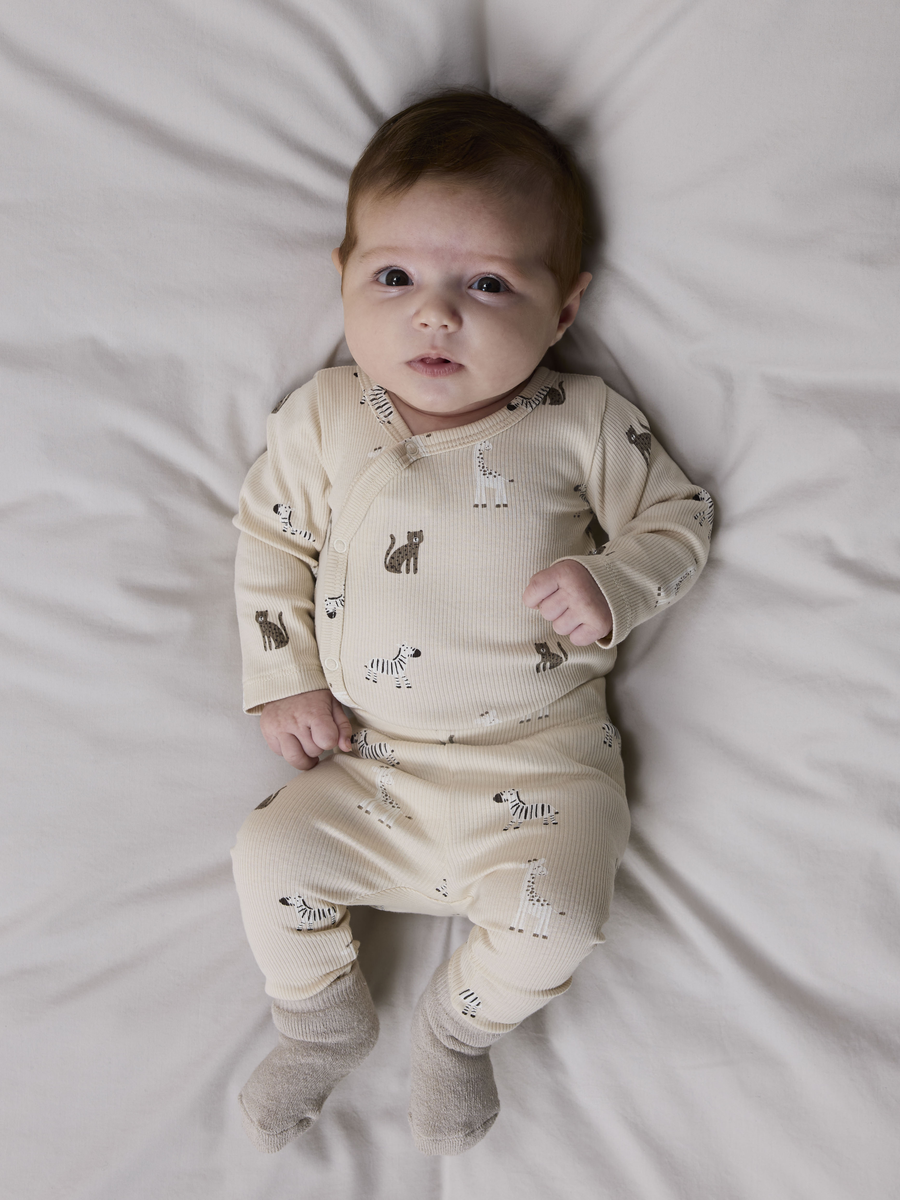 Baby har på seg en lys beige ribbet heldress med dyremønster og omslagsknapping samt lysegrå sokker.