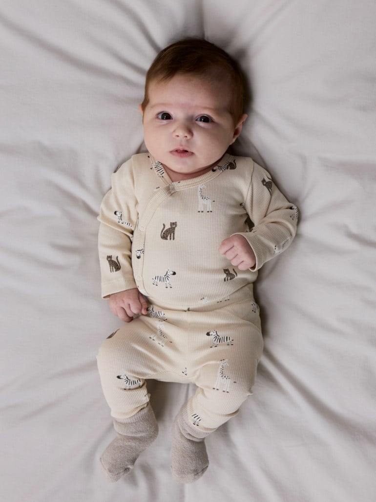 Baby har på seg en lys beige ribbet heldress med dyremønster og omslagsknapping samt lysegrå sokker.