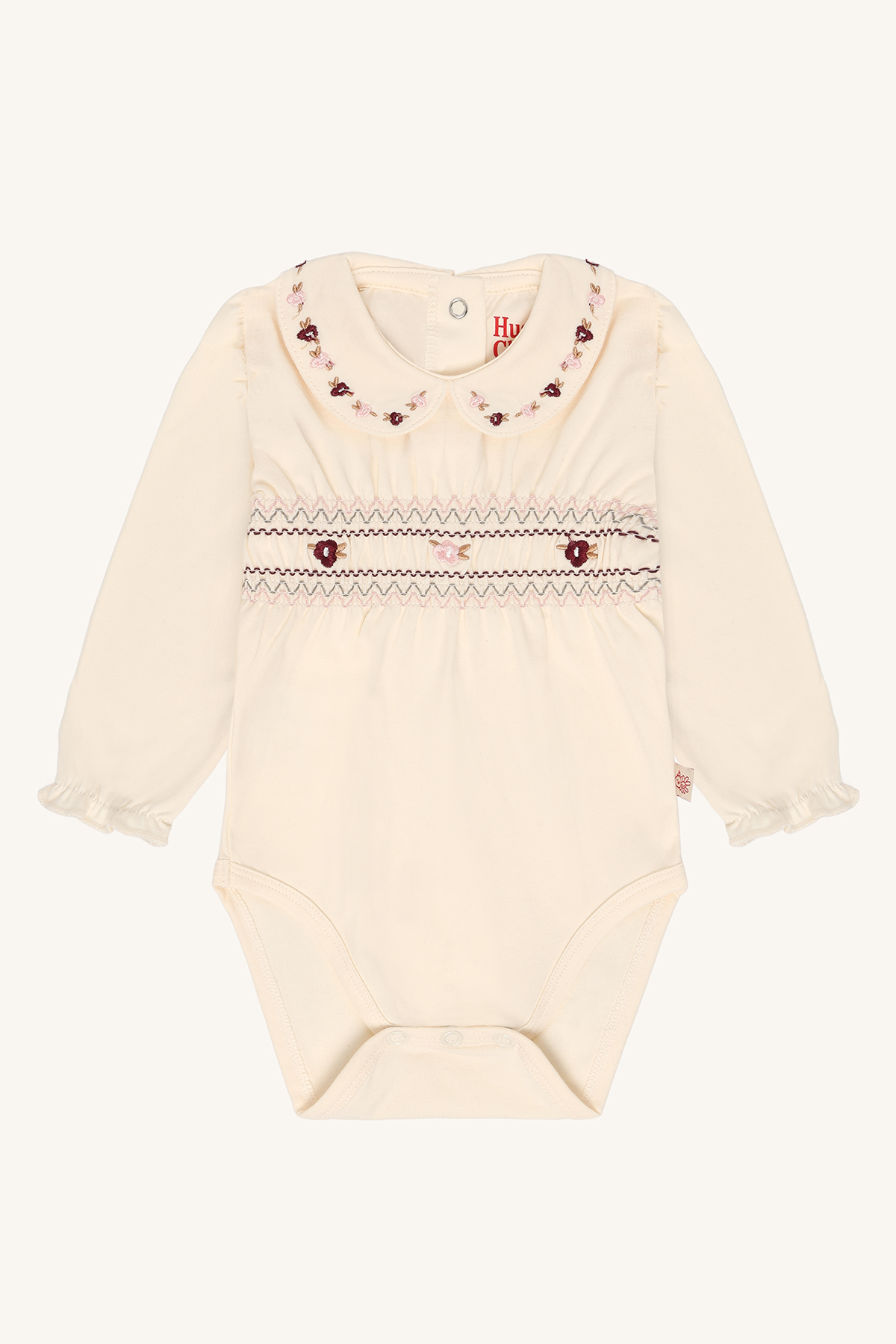 En kremfarget babybody med lange ermer, rund brodert blomsterkrage og smockedetaljer på brystet.