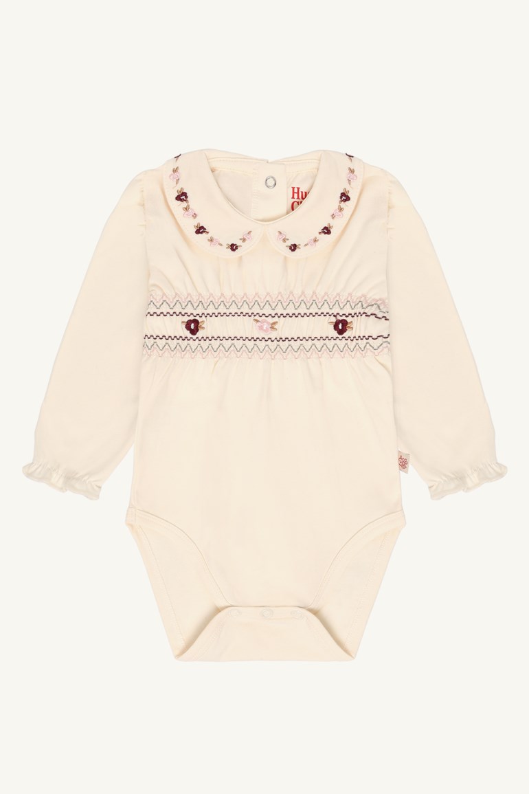 En kremfarget babybody med lange ermer, rund brodert blomsterkrage og smockedetaljer på brystet.