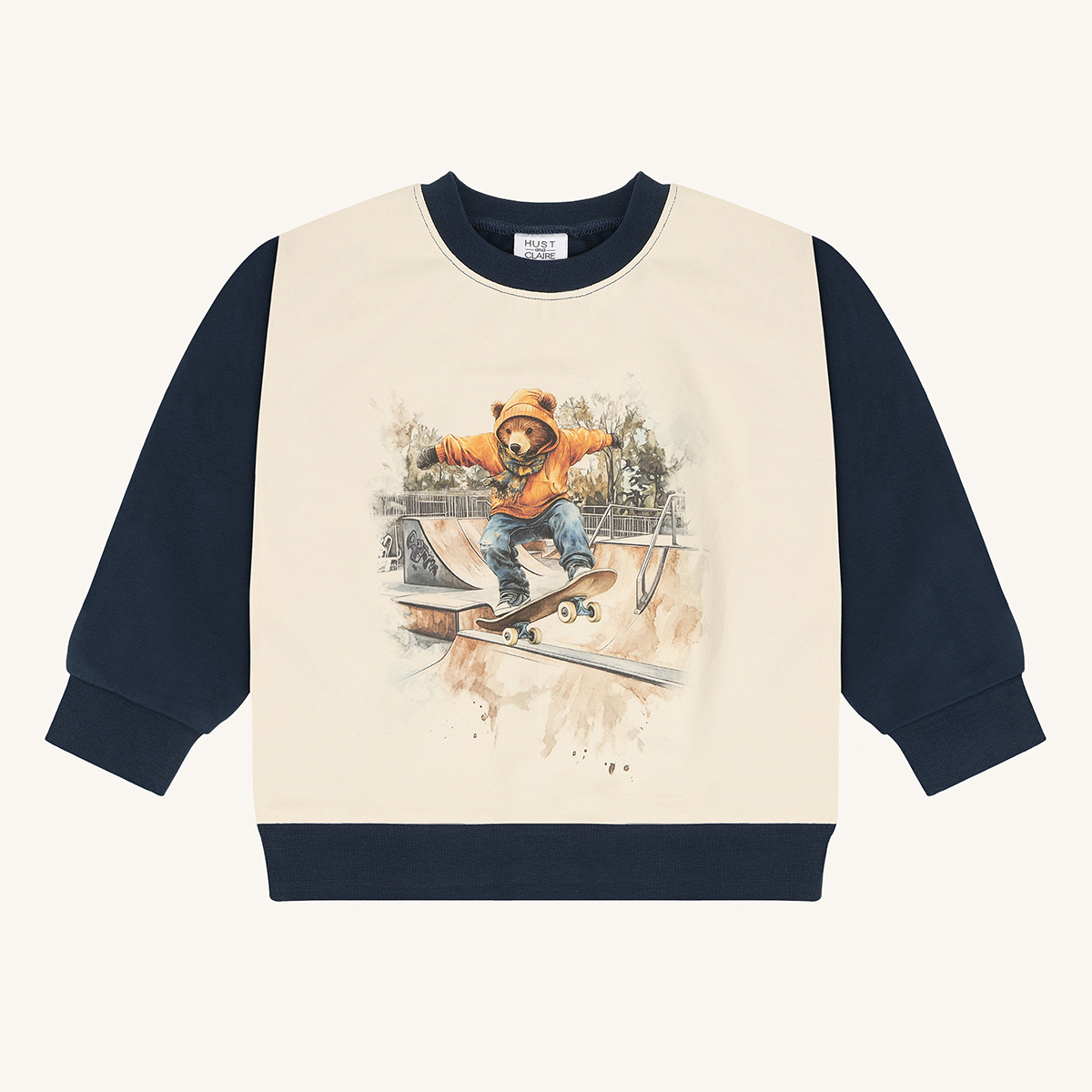 Langermet barnsgenser med kremfarget frontpanel, marineblå ermer og et trykk av en skateboardkjørende bjørn.