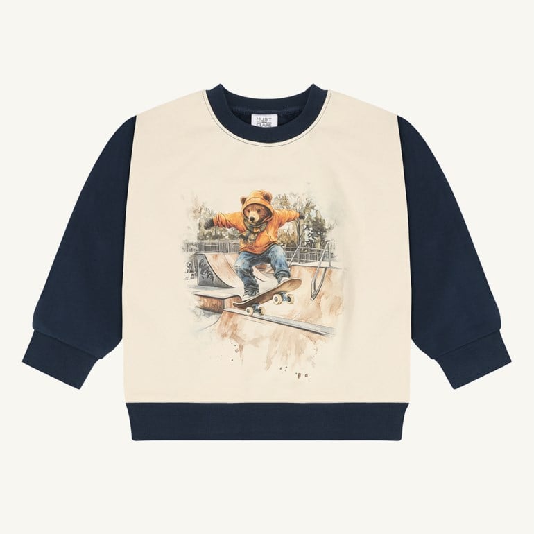 Langermet barnsgenser med kremfarget frontpanel, marineblå ermer og et trykk av en skateboardkjørende bjørn.