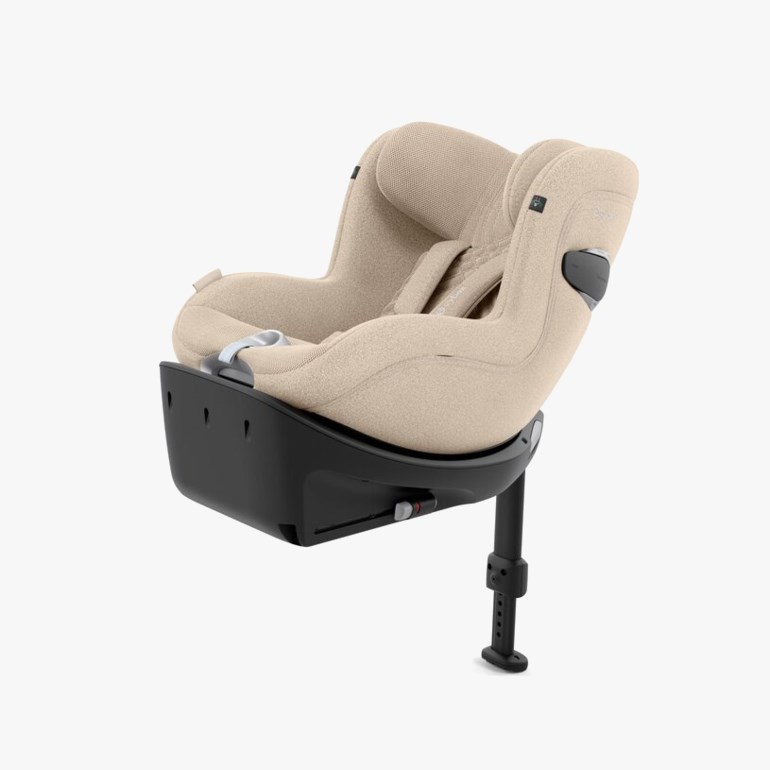 Beige babybilsete i teksturert stoff på svart roterende base med støtteben, skrått forfra.