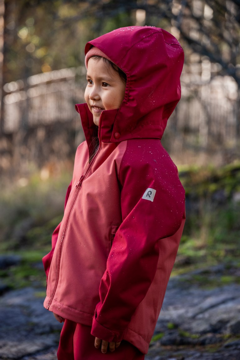 Barn med tofarget rød vanntett jakke med hette, glidelås, 'R'-logo, rosa lue og rød bukse.