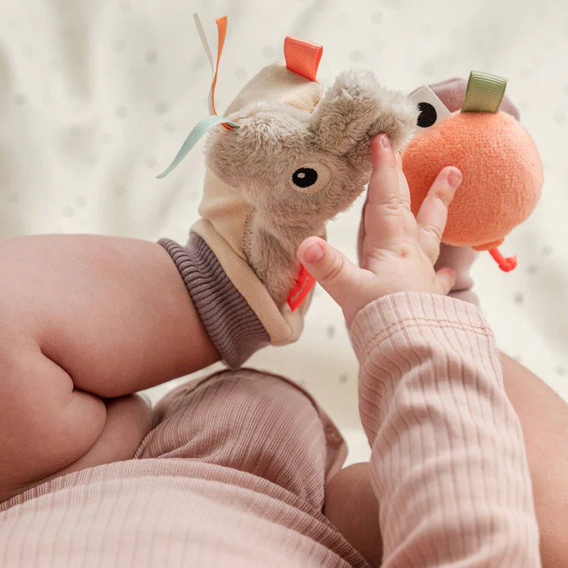 Baby har på beige dyreformet håndleddsrangl med brun ribb, berører oransje rangl, og bærer lys rosa topp.