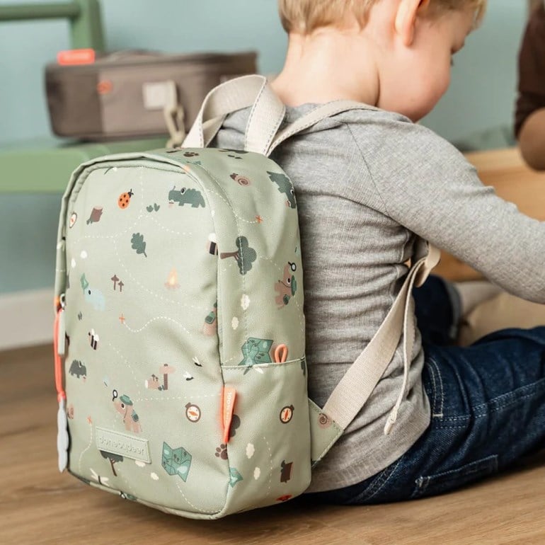 Et barn bærer en lysegrønn mønstret ryggsekk over en grå langermet topp og blå jeans, sittende på et gulv.
