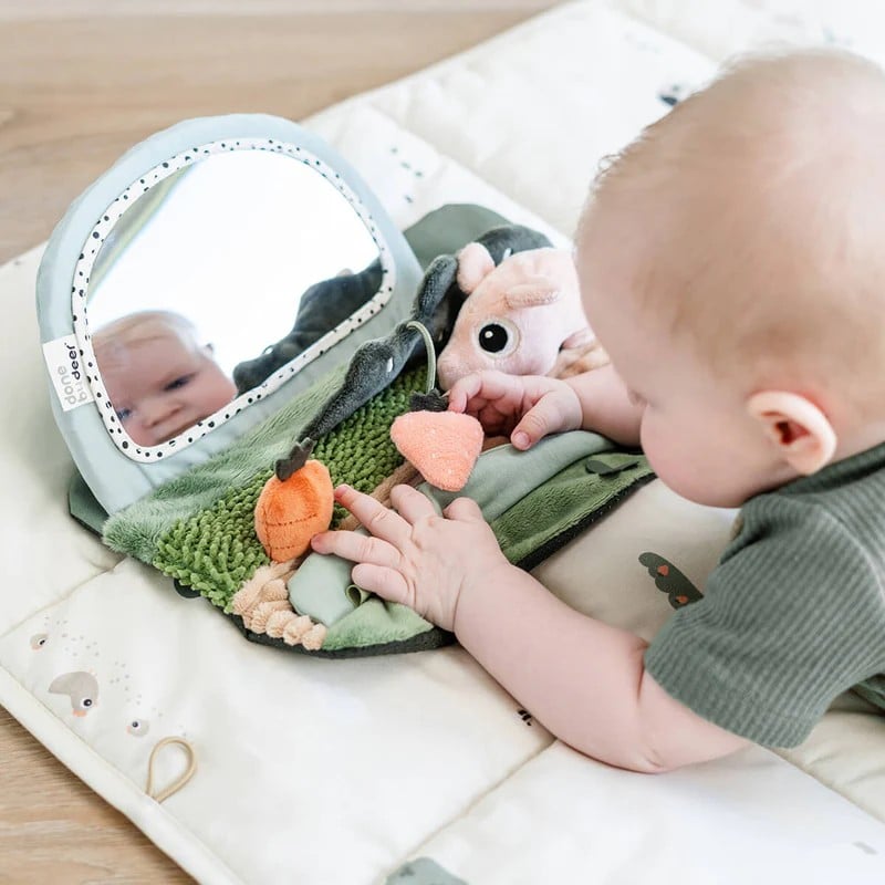 Baby på en aktivitetsmatte med speil, plysjgulrøtter og et mykt dyr, iført en grønn ribbet topp.
