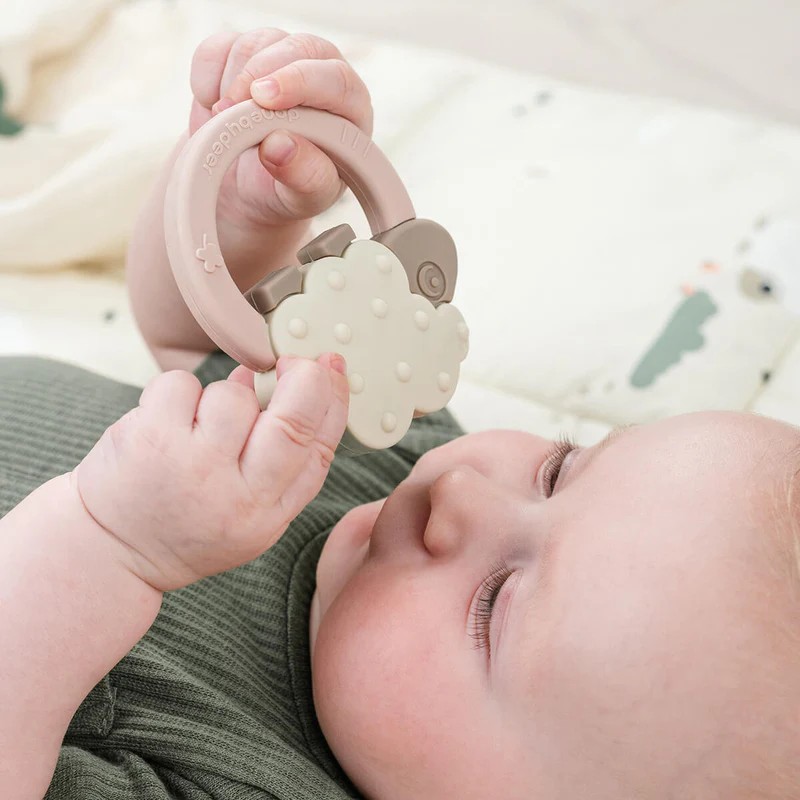Baby holder en rosa og beige skyformet silikongrepeteether, iført en grønn ribbet topp.