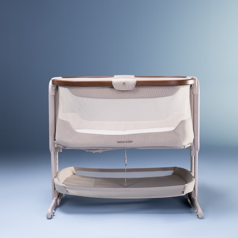 En lys beige babyseng med nettingvegger, en topplist i tre og en nedre oppbevaringskurv.