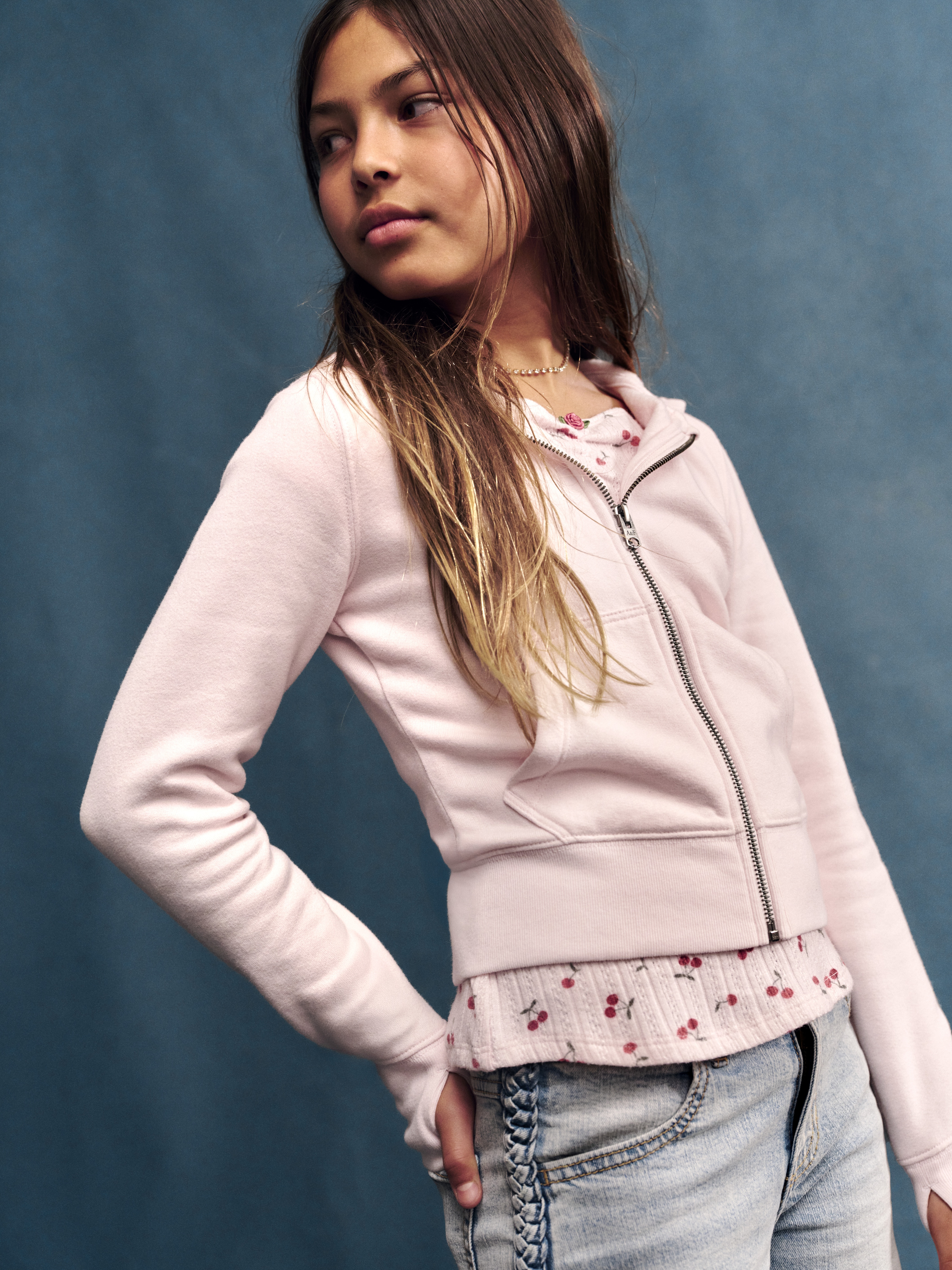 Barn i lys rosa hettegenser med glidelås over lys rosa topp med kirsebærmønster og lys blå jeans.