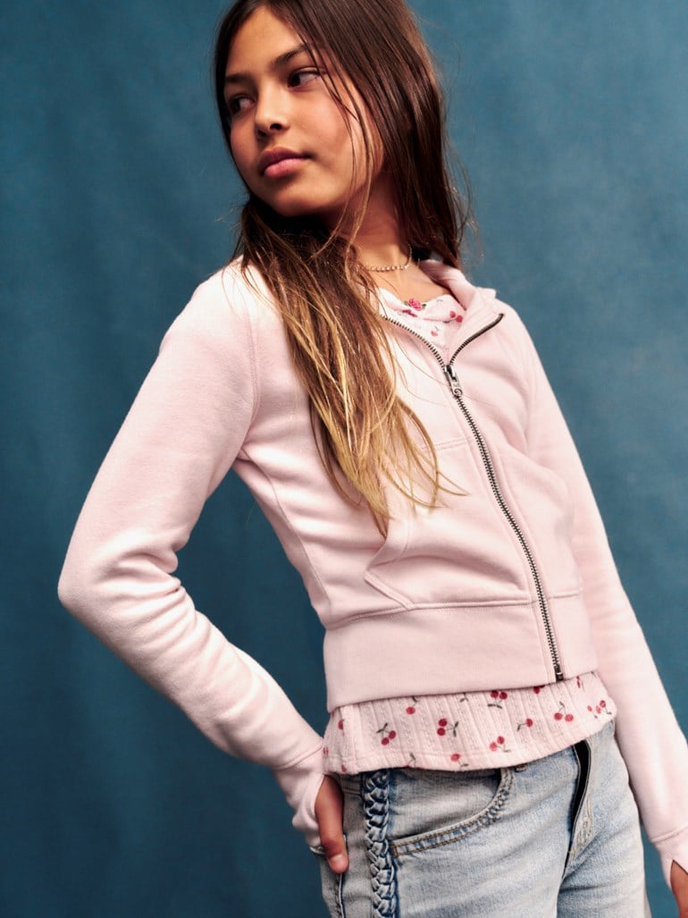 Barn i lys rosa hettegenser med glidelås over lys rosa topp med kirsebærmønster og lys blå jeans.