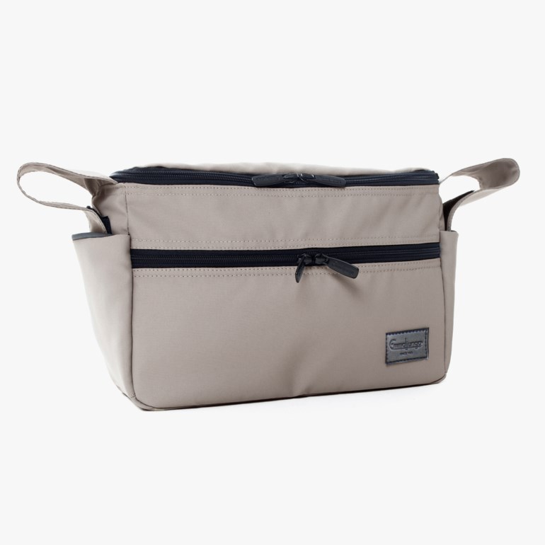 Lys beige veske med svarte glidelåser, en frontlomme og sidehåndtak, forfra.
