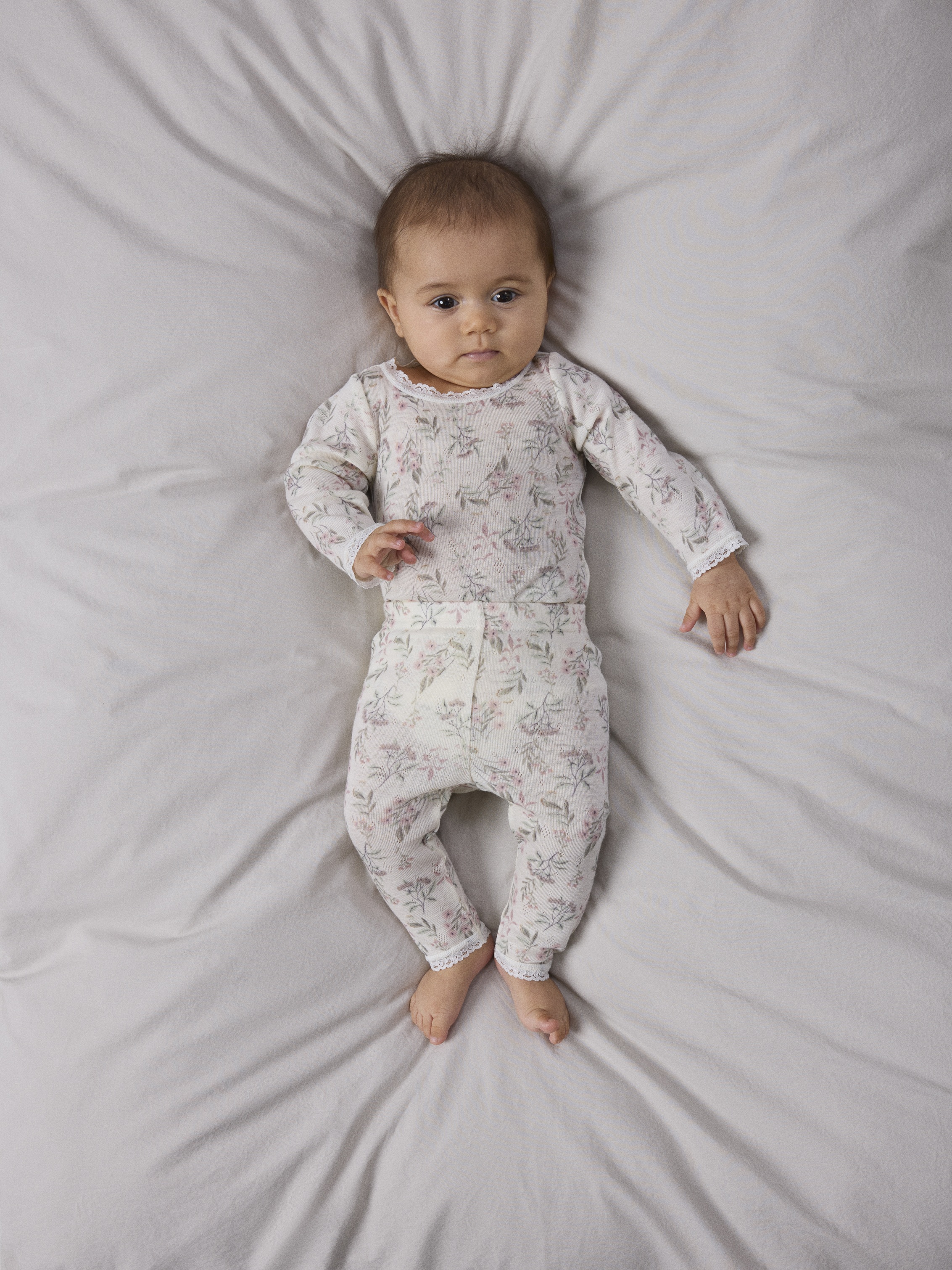 Baby har på seg et todelt langermet lyst blomstret pysjamassett med blondekant.