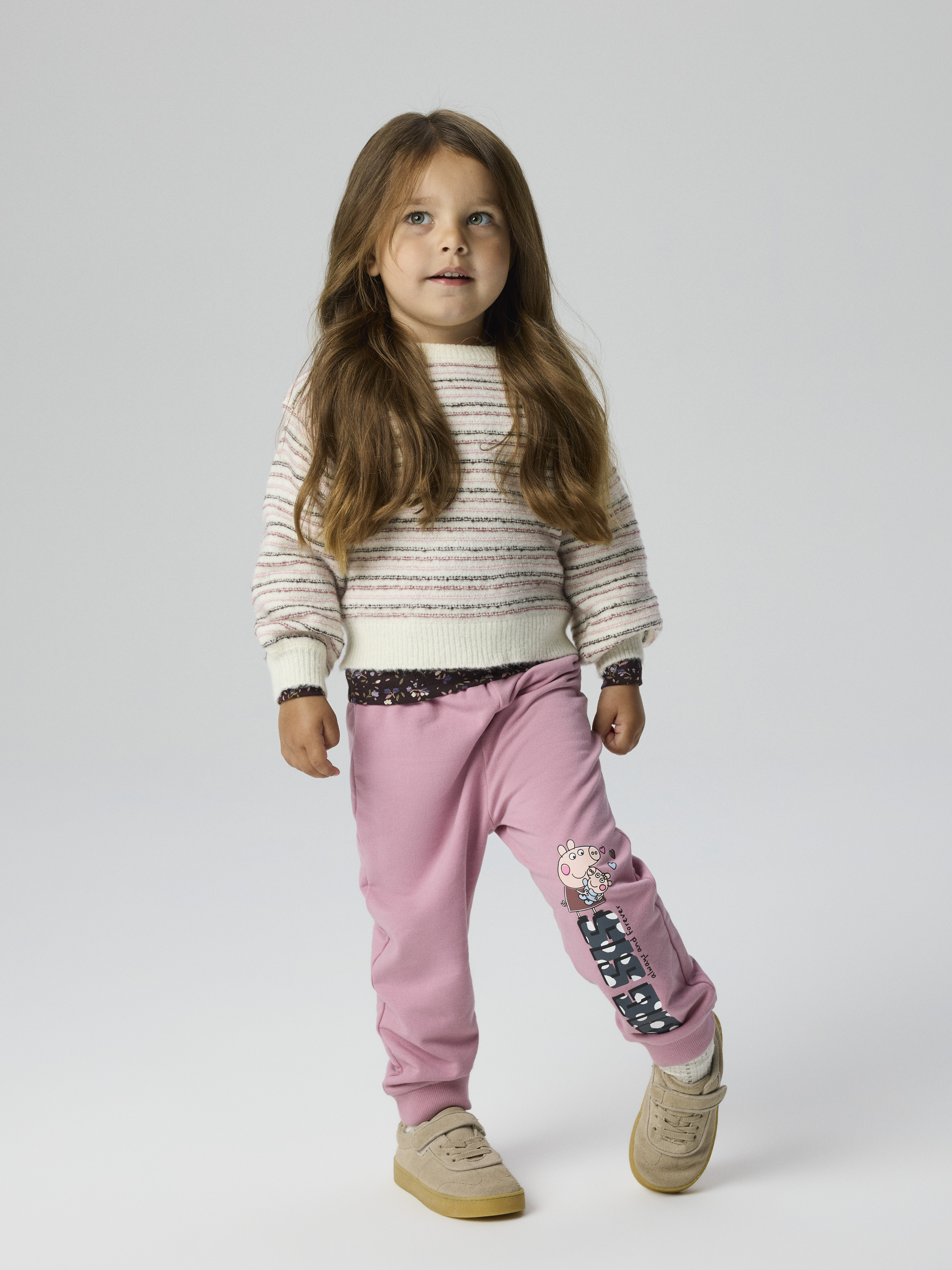 Barn i rosa joggebukse med figurtrykk, stripet strikkegenser, mønstret topp og brune joggesko.