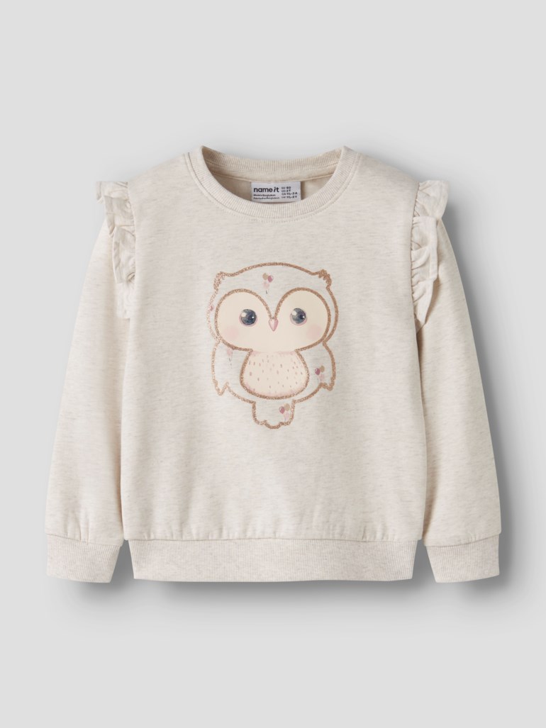 Barnegenser i lys beige melert med rysjedetaljer på skuldrene, ribbestrikkede mansjetter og et glitrende ugletrykk.