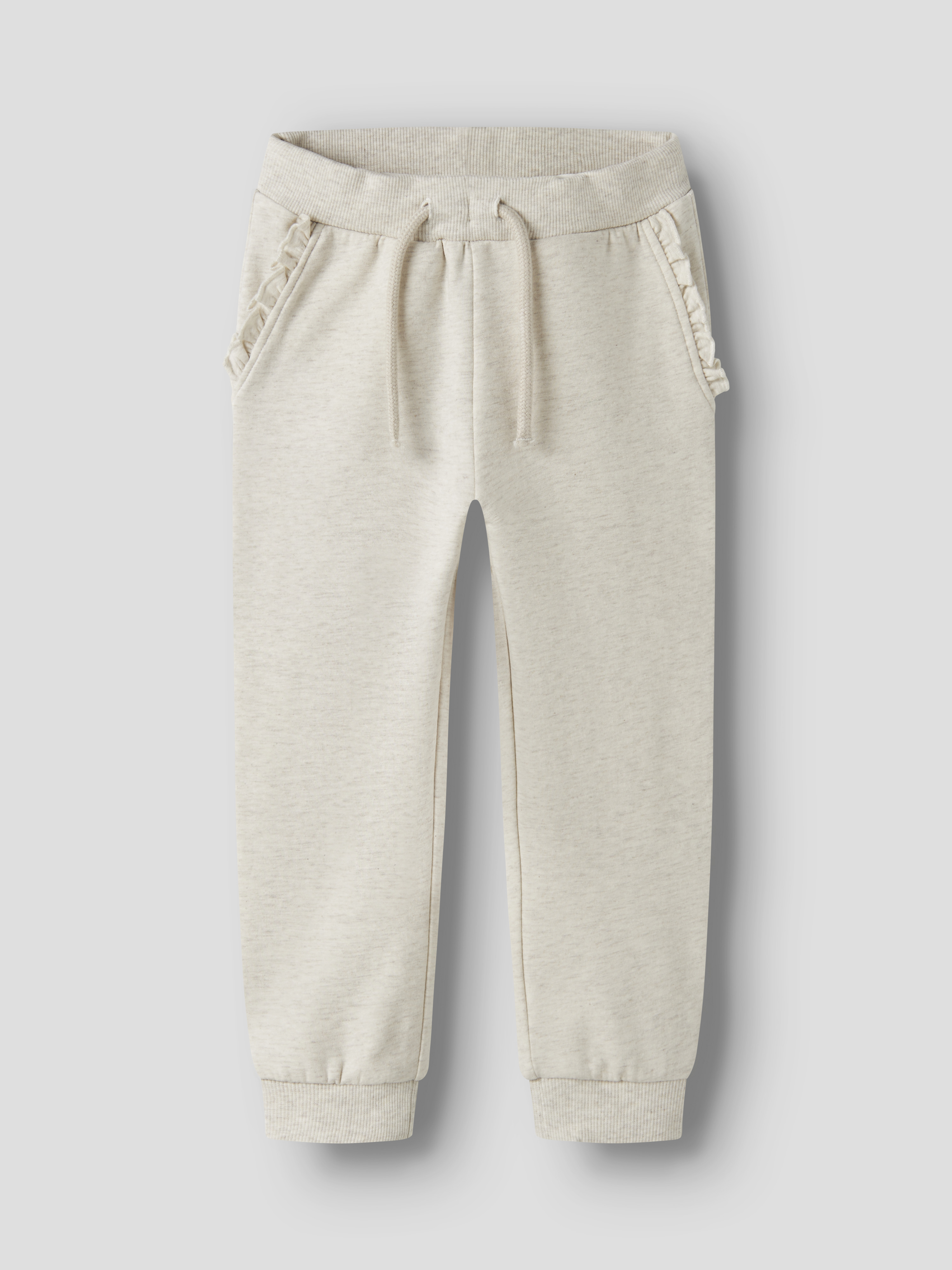 Lys beige barne-joggebukse med snøring i midjen, volangprydde sidelommer og ribbestrikkede mansjetter, forfra.