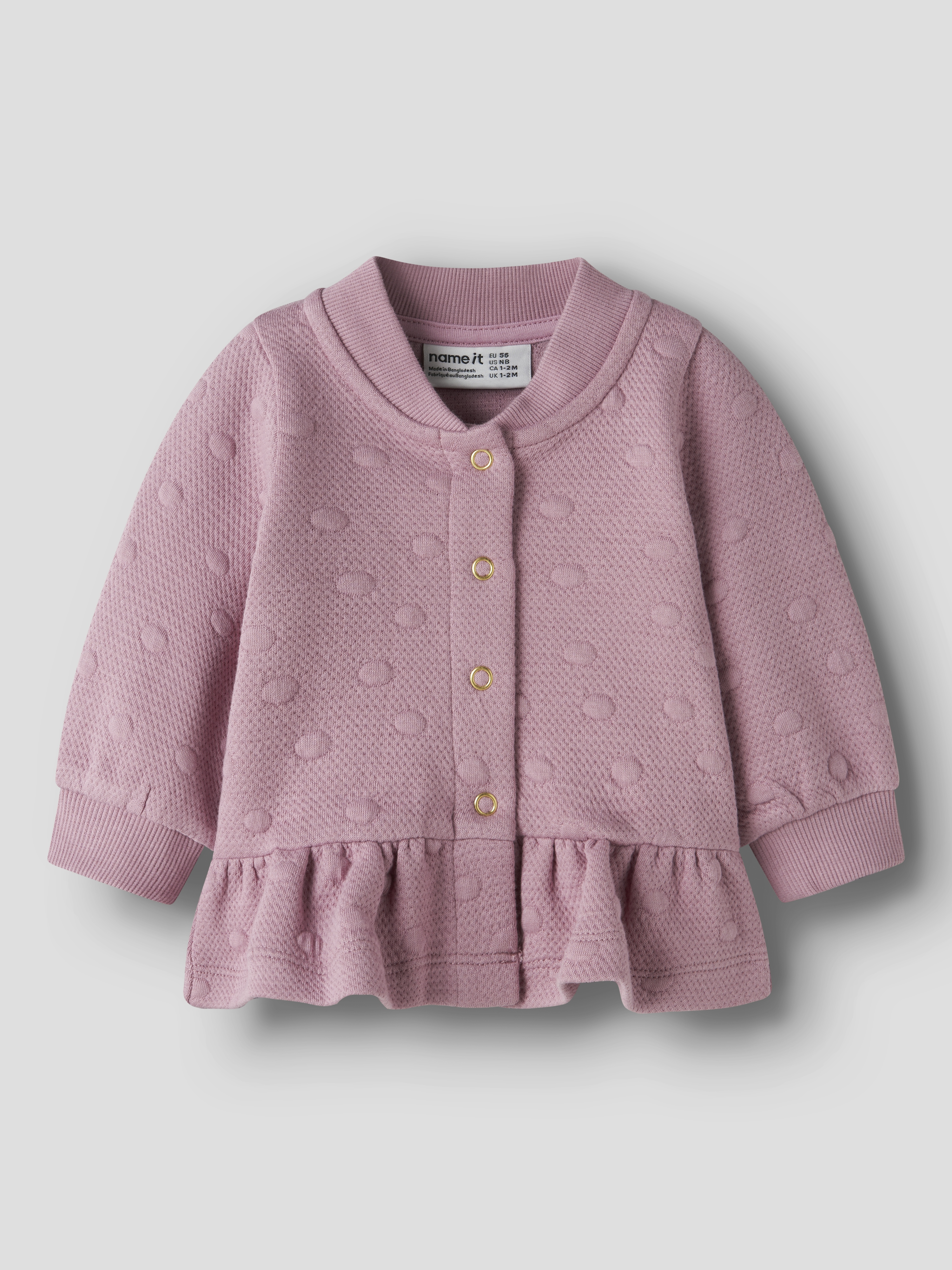 Forfra, en rosa babycardigan med prikket mønster, gullfargede knapper og volang nederst.
