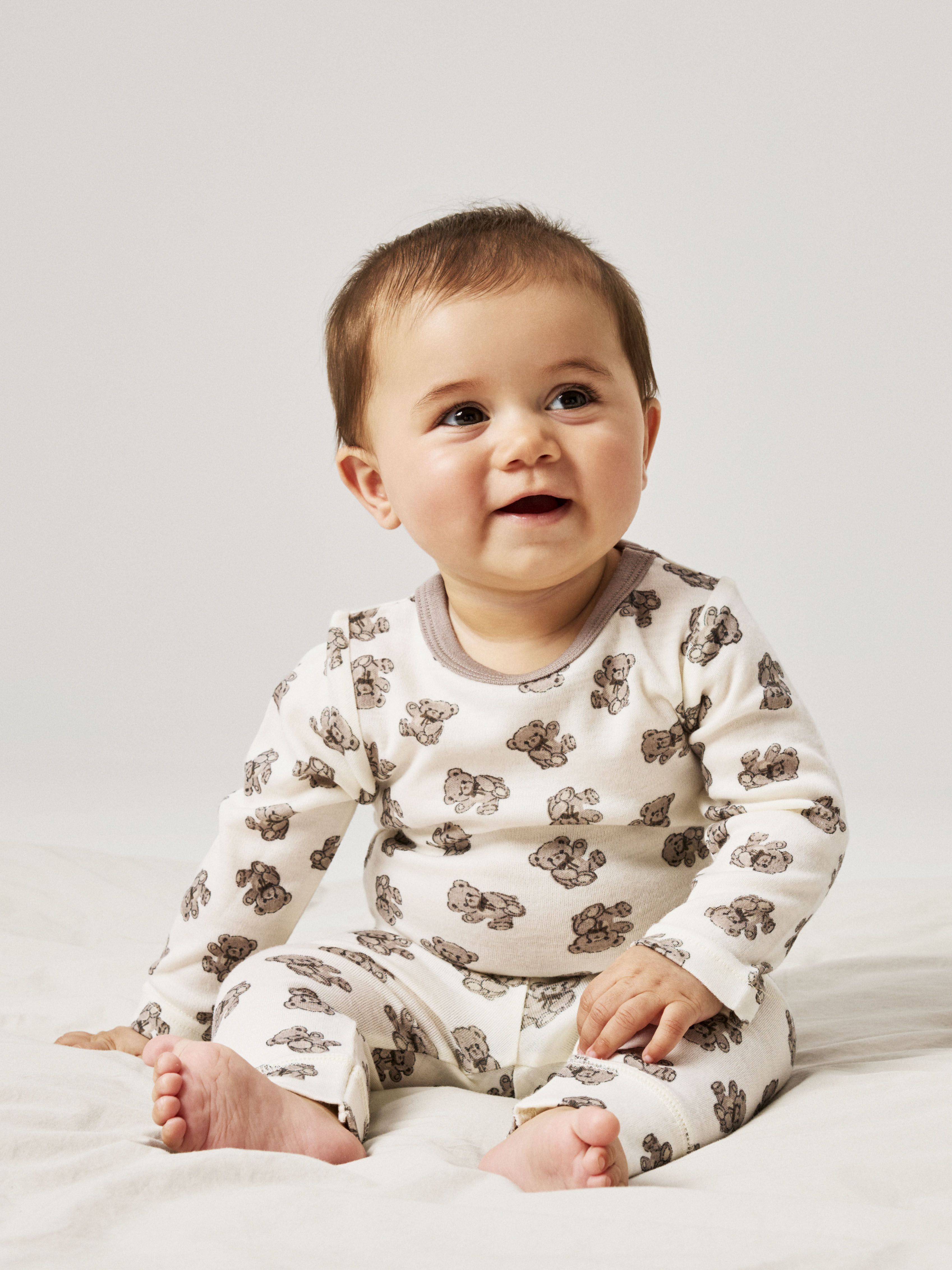 Baby i et kremfarget langermet todelt sett med brunt teddybjørnmønster og brun halskant.