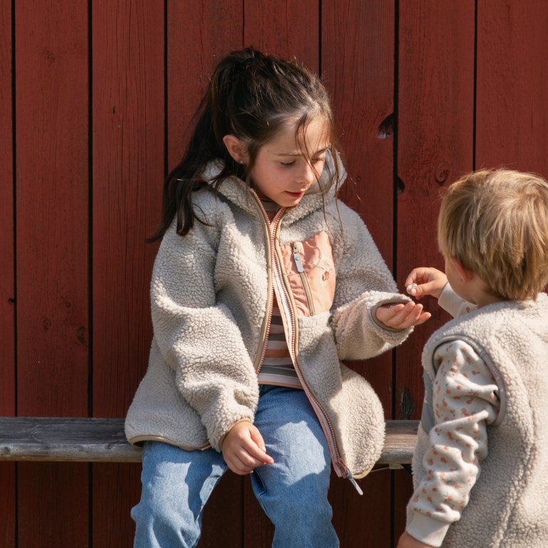 Barn i beige teddyfleecejakke med rosa glidelås mottar gjenstand fra barn i matchende vest.