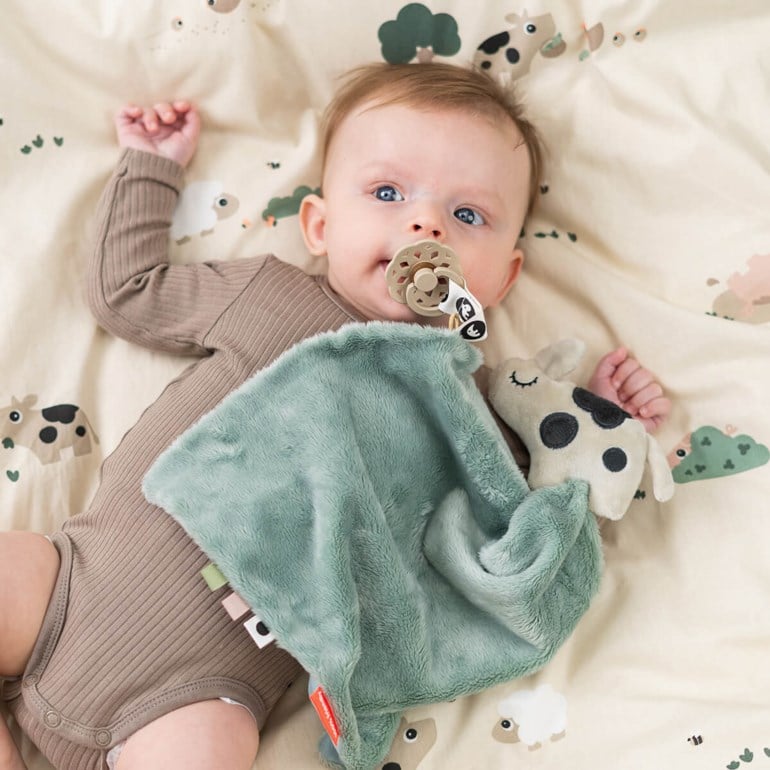 Baby holder salviegrønt plysj-kosepledd, iført brun ribbet body, med beige smokk og flekkete ku-leketøy.