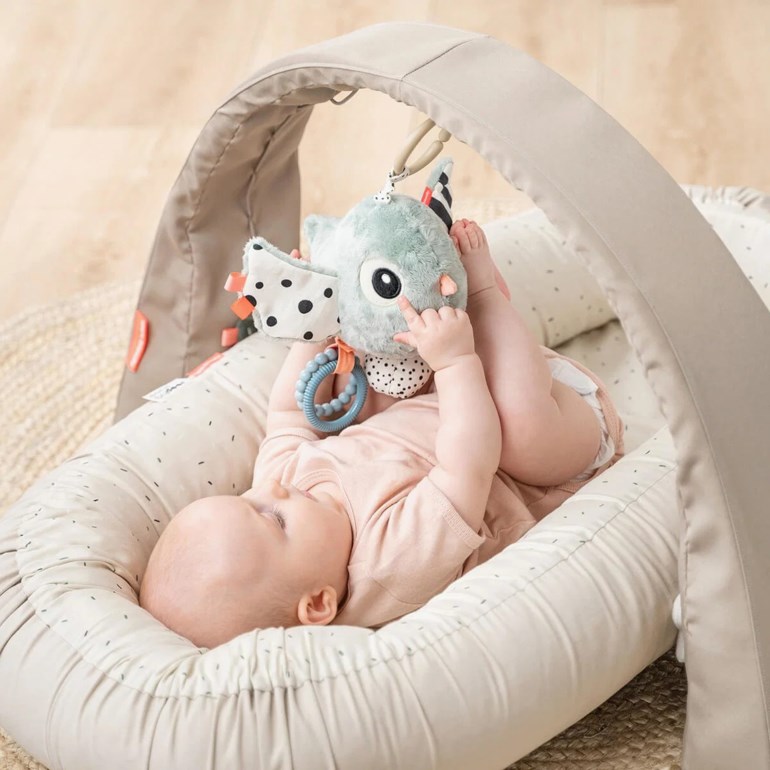 Baby ligger i et beige aktivitetsgym med en bue og leker med en mintgrønn plysj dinosaurleke.