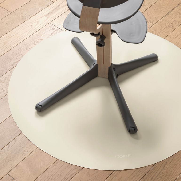 En rund, lys beige gulvbeskyttelsesmatte under en grå og trefarget barnestol med kryssfott.