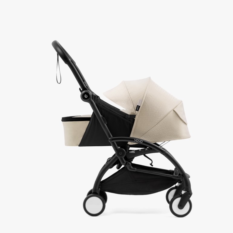 Sideprofil av en svart barnevogn med en beige mønstret kalesje, en svart og beige handlekurv og hvite hjul.