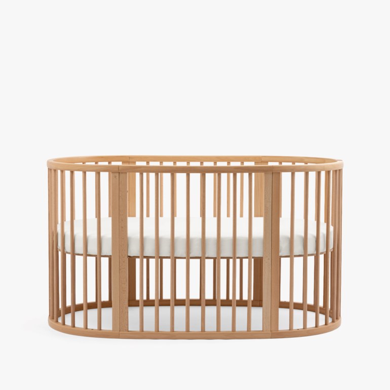Lys trefarget oval babyseng med treribber og hvit madrass.